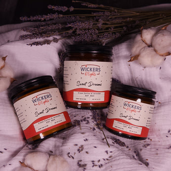 A serene setup of three Wickers D'Lights Sweet Dreams soy wax jars with a clean cotton and lavender scent, arranged on a soft fabric adorned with lavender sprigs and cotton bolls, evoking a soothing and calm atmosphere.