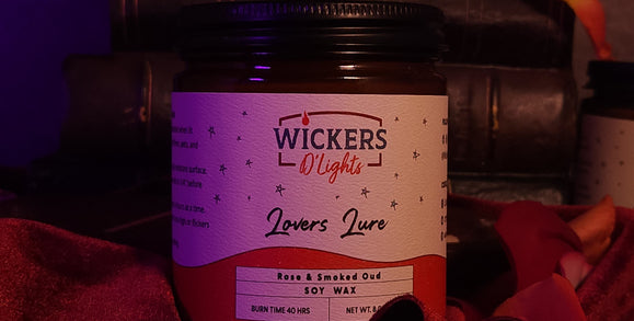 A romantic and moody setting featuring a jar of Wickers D'Lights Lovers Lure soy wax with a rose and smoked oud scent, accentuated by soft lighting, velvety fabric, and scattered rose petals, with a dark red rose in the blurred background.