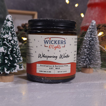 A Wickers D'Lights Whispering Winter soy wax candle, with a net weight of 3.8 oz and a burn time of 20 hours, is displayed. The candle's label features the Wickers D'Lights logo and the scent notes of Eucalyptus & Rainwashed Pine. It is presented against a cozy, festive backdrop with miniature snowy Christmas trees, soft lighting, and holiday decorations.