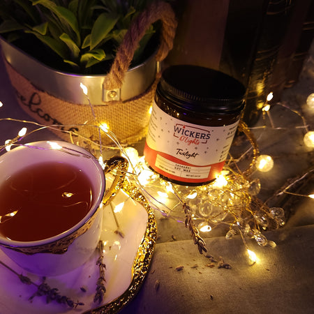 A warm and inviting scene featuring a jar of Wickers D'Lights Twilight soy wax, a cup of tea beside it on an ornate saucer, all bathed in the glow of fairy lights. Dried lavender and a potted green plant in the background add to the tranquil, evening mood.