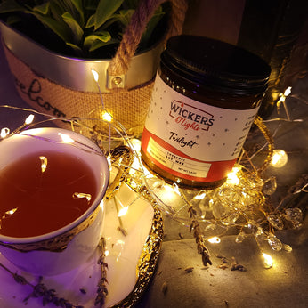 A cozy scene with a jar of Wickers D'Lights Twilight soy wax next to a cup of tea on a saucer, surrounded by fairy lights, with lavender sprigs and a potted plant in the background, creating an inviting and relaxed ambiance.