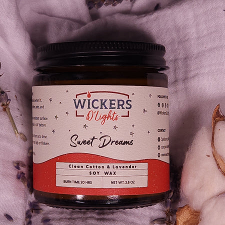 Centered in the frame is a Wickers D'Lights Sweet Dreams soy wax jar, with a label indicating the clean cotton and lavender scent. The jar is nestled amidst a soft, white fabric with scattered lavender flowers and cotton bolls, evoking a sense of calm and comfort.