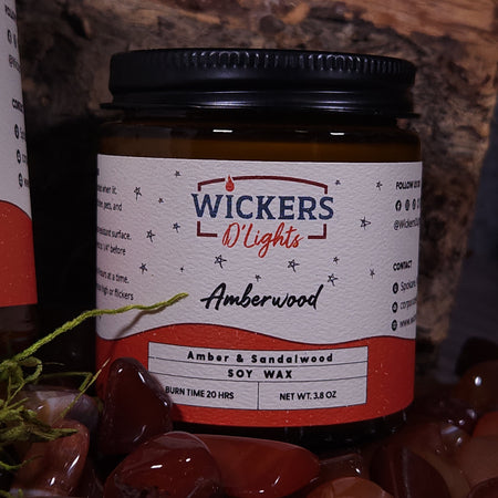 A close-up of a Wickers D'Lights soy wax candle in the 'Amberwood' scent. The candle is set against a rustic backdrop, with polished amber stones in the foreground. The label on the amber-colored jar with a black lid details the scent as Amber & Sandalwood, a burn time of 20 hours, and a net weight of 3.8 oz. The label design features decorative stars and the company's branding in a clean, elegant font.